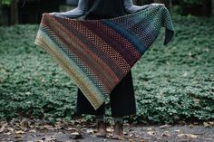 a woman holding up a multicolored shawl in front of some bushes and trees