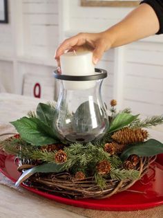 a person is pouring white candle into a glass vase on a red plate with pine cones and greenery