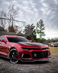 a red chevrolet camaro is parked in front of a helicopter