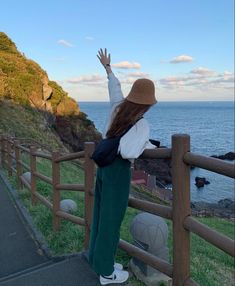 a woman standing on top of a lush green hillside next to the ocean with her arms in the air