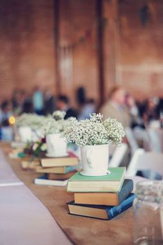 several books are lined up on a table with flowers in them and people sitting at tables behind them