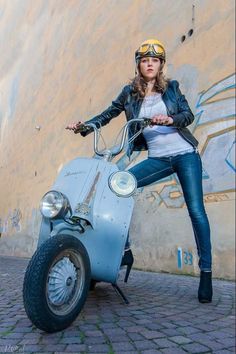 a woman in leather jacket and goggles sitting on an old scooter by a wall