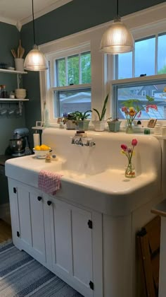 a white sink sitting under two windows in a kitchen next to a counter with flowers on it