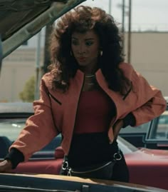 a woman standing in front of a car with her hand on the hood and looking at the camera