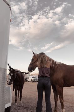 a woman standing next to a brown horse on top of a dirt field near a trailer