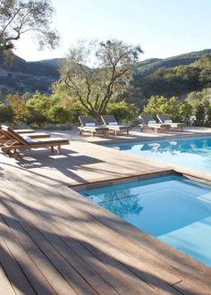 a wooden deck next to a swimming pool with chaise lounges and trees in the background