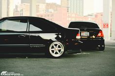 two black cars parked next to each other in a parking lot with tall buildings behind them