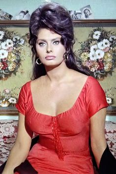 a woman in a red dress is sitting on a couch with flowers behind her and looking at the camera