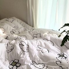 an unmade bed with white sheets and black drawings on it, next to a plant