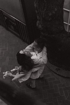 black and white photograph of two people hugging each other on the sidewalk near a tree