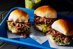 three pulled pork sandwiches with guacamole sauce on a blue tray next to a green drink