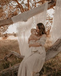 a woman holding a baby in her arms while sitting on a tree branch with white fabric hanging from the branches