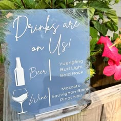 a sign that says drinks are on us and wine is in front of some flowers