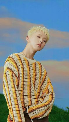 a young man with blonde hair wearing a yellow and white knitted sweater standing in front of a blue sky