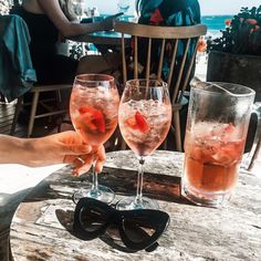 two people are holding up their glasses with some fruit in them on a table by the beach