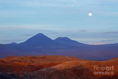 the moon is setting over some mountains in the desert with orange rocks and grass on the ground