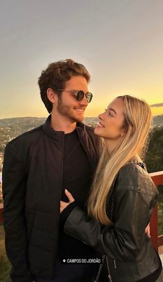 a man and woman standing next to each other in front of the sun setting on top of a hill