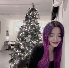 a woman with long purple hair standing next to a christmas tree