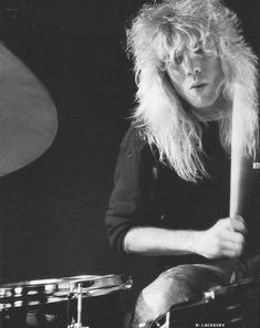 a man with long hair sitting in front of a drum set and holding a stick