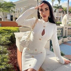 a woman is posing for the camera wearing a white dress and gold jewelry, with palm trees in the background