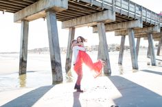 a woman in an orange dress is dancing on the beach under a pier with her legs spread out