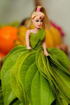 a doll in a green dress sitting on top of a leafy plant with oranges behind it