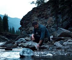 a man crouches on rocks in the middle of a stream as he looks at his cell phone