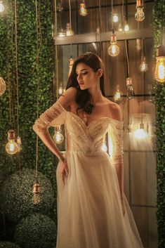 a woman in a wedding dress standing next to a wall with hanging lights and plants