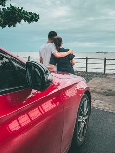 two people sitting on the hood of a red sports car looking out at the ocean