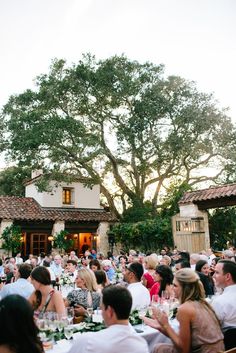 a large group of people sitting at tables in front of a house with trees and bushes