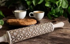 a cookie on a plate next to two cups of coffee and a rolling pin that is shaped like a flower