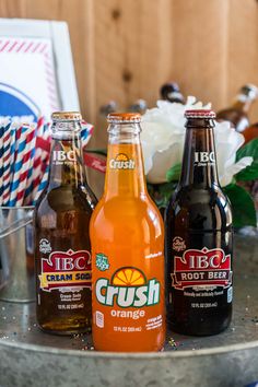 three bottles of orange soda sitting on top of a metal table next to a flower