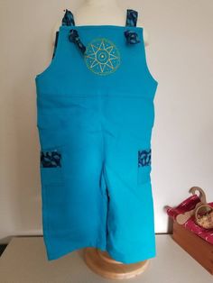 Size 5 unisex overalls. Made of a sturdy, yet soft, cotton pique in teal blue. Contrast straps and side pockets feature a.dark blue cotton batik. Hand embroidered sunburst on the front bib. Sleeveless Blue Cotton Overalls, Cotton Overalls With Side Pockets, Cotton Overalls For Summer Festivals, Cotton Summer Overalls For Festivals, Summer Festival Cotton Overalls, Casual Cotton Overalls For Festivals, Blue Cotton Overalls With Pockets, Cotton Overalls With Pockets For Festival, Casual Embroidered Cotton Overalls