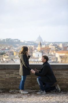 a man kneeling down next to a woman on top of a brick wall with a city in the background