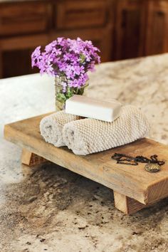 a wooden tray with soap and towels on it sitting on a marble counter top next to a vase filled with purple flowers