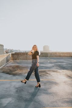 a woman in black shirt and jeans walking across an empty parking lot with buildings in the background