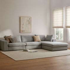 a living room with white walls and wooden flooring, large sectional couch in front of two windows