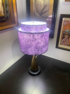 a purple lamp shade sitting on top of a wooden table next to pictures and paintings