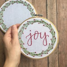 someone is holding up two embroidery hoops with the word joy on them, which are decorated in red and green