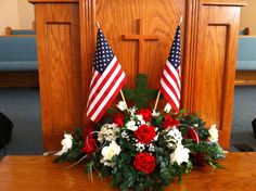 two american flags and flowers sit on a table in front of a wooden wall with pews