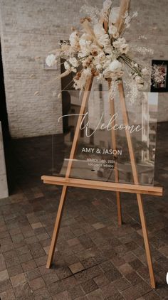 a wooden easel with flowers on it and the words welcome are displayed in front of a brick wall