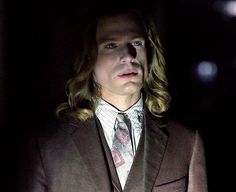 a man with long hair wearing a suit and tie standing in the dark, looking to his left