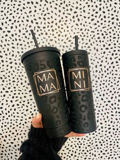 two black tumblers with the words mam on them are held in front of a leopard print wall