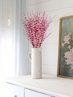 a white vase with pink flowers sitting on top of a table next to a painting