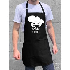 a man wearing an apron with the words best chef in white on it, standing next to a brick wall