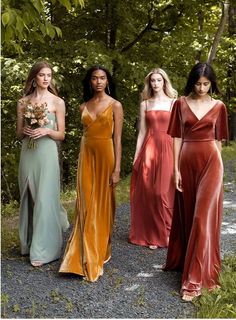 four women in long dresses standing on a gravel road with trees and bushes behind them