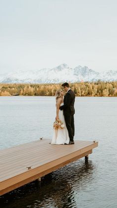 a bride and groom standing on a dock in front of the water with mountains behind them