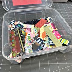 a plastic container filled with lots of different types of fabric and paper pieces on top of a tiled floor