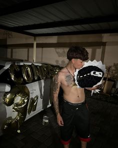 a shirtless man holding a black object in front of a white car with gold letters on it