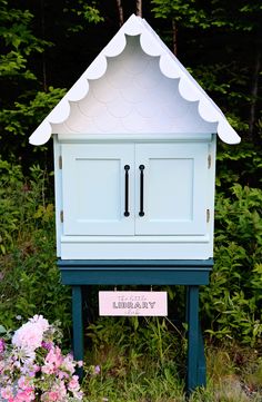 a blue birdhouse with a white roof and two doors on the front is surrounded by pink flowers